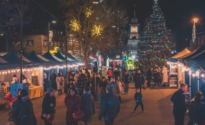 A festive Christmas market at night, with shoppers strolling between stalls adorned with string lights. A large, decorated tree and clock tower glow in the background.