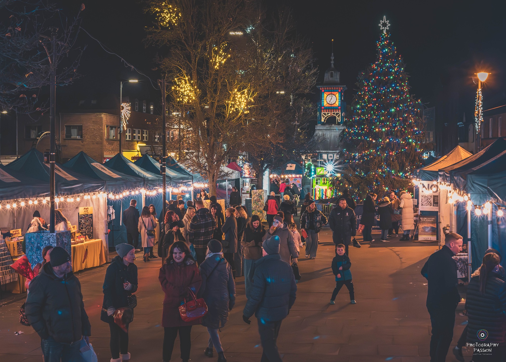 A festive Christmas market at night, with shoppers strolling between stalls adorned with string lights. A large, decorated tree and clock tower glow in the background.