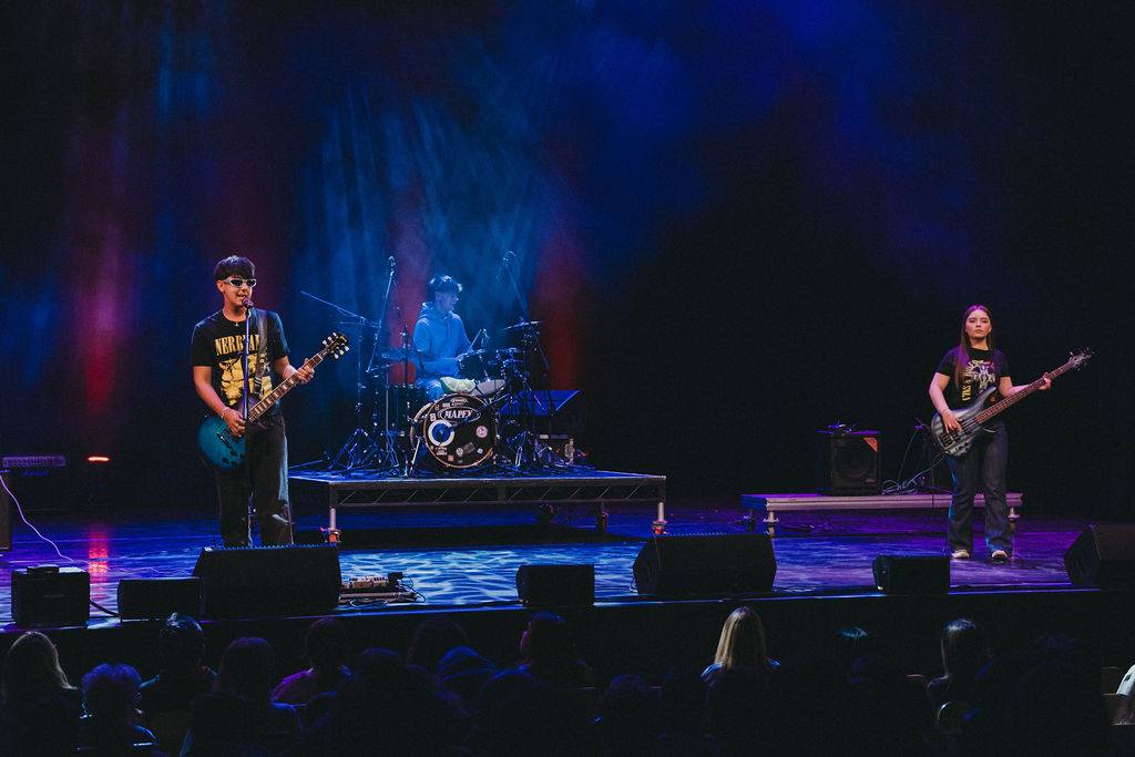 A three-piece rock band performs on stage under vibrant red and blue lights. The scene conveys an energetic and lively atmosphere, with a guitarist and bassist flanking a drummer.
