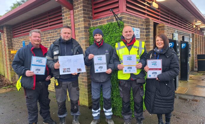Five people stand smiling in front of a brick building, holding certificates. They wear winter clothing, including jackets and a high-visibility vest.