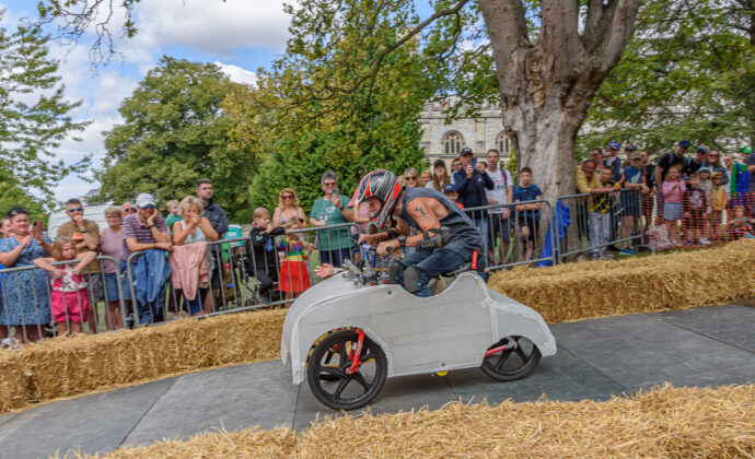 A racer in a small, white soapbox car speeds along a track lined with hay bales. Spectators watch eagerly from behind a fence, under clear skies.