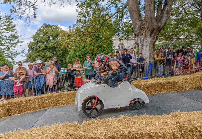 A racer in a small, white soapbox car speeds along a track lined with hay bales. Spectators watch eagerly from behind a fence, under clear skies.