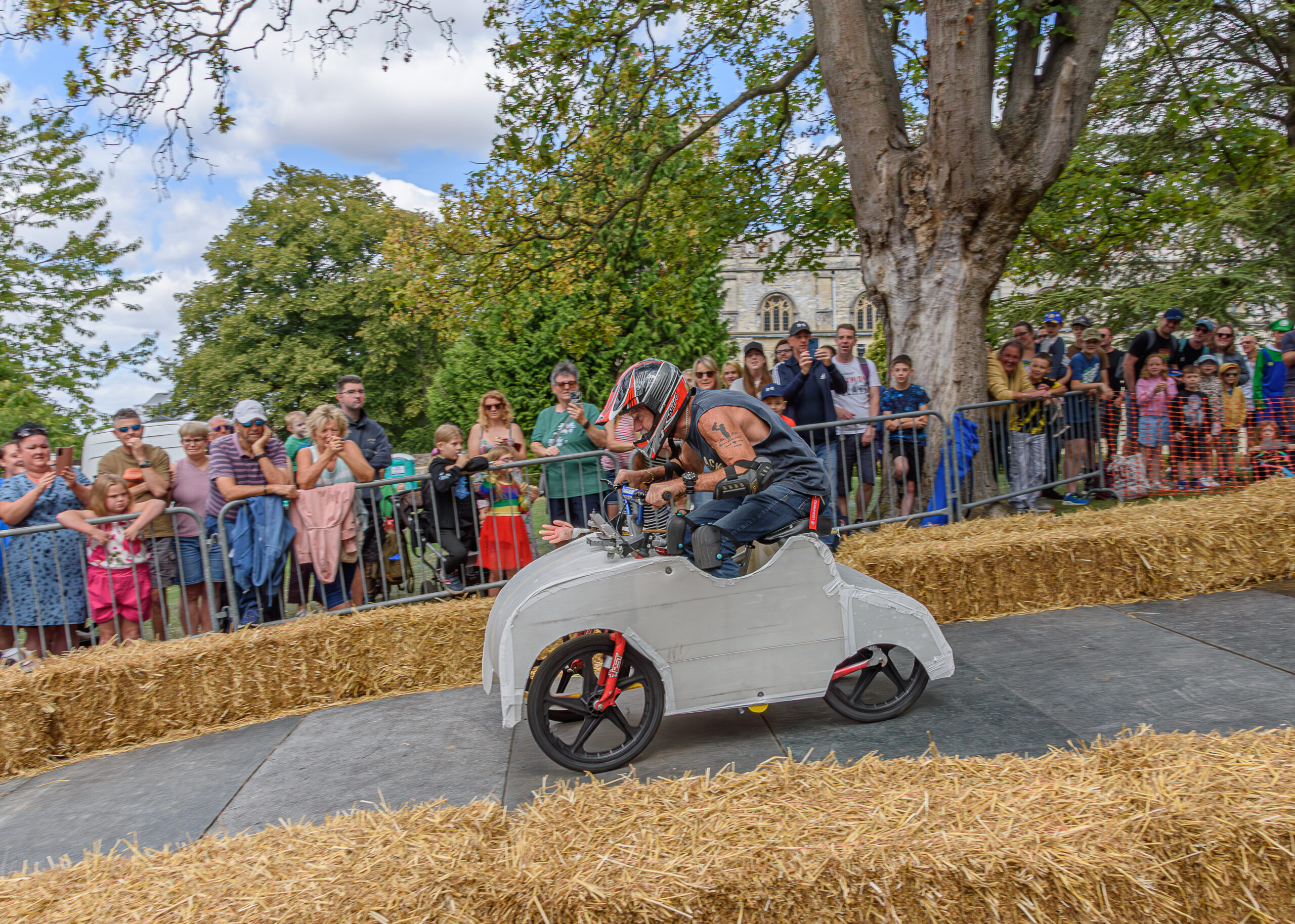 A racer in a small, white soapbox car speeds along a track lined with hay bales. Spectators watch eagerly from behind a fence, under clear skies.