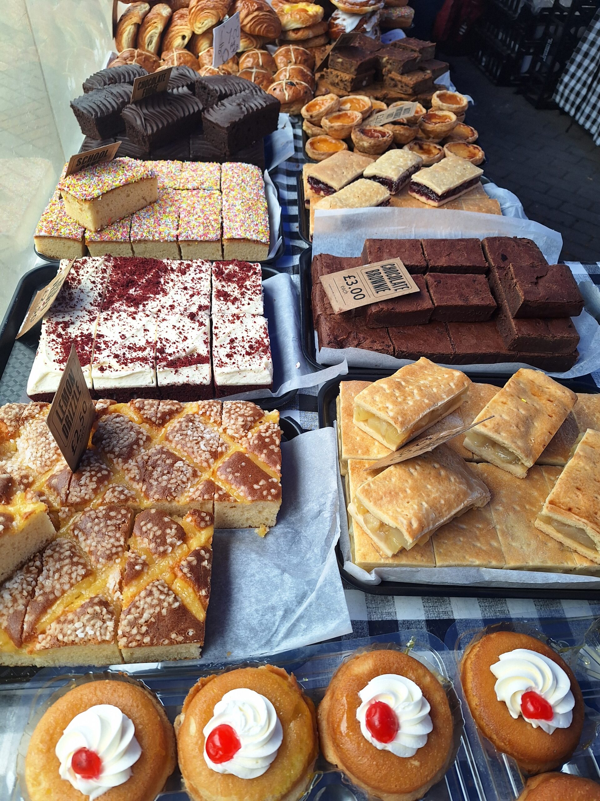 An array of assorted pastries on a display table, including layered cakes, red velvet slices, pastries with glaze, and cupcakes topped with cherries, convey a tempting and indulgent atmosphere. Bread on the Rise at Middle Row markets