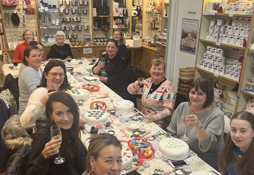 A group of joyful women sit around a table in a craft store, holding drinks and laughing, surrounded by decorated cakes and colourful crafting materials.
