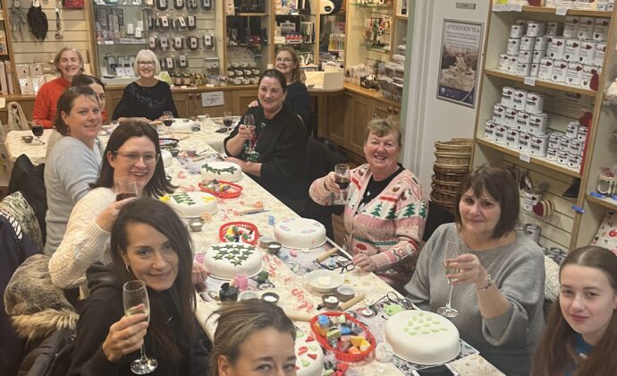 A group of joyful women sit around a table in a craft store, holding drinks and laughing, surrounded by decorated cakes and colourful crafting materials.