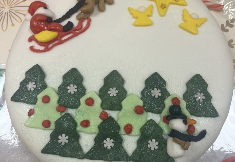 Round cake decorated with a festive scene: Santa in a sleigh, stars, and angels in the sky, evergreen trees with snowflakes, and a snowman below.