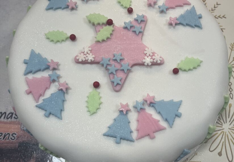 Festive cake with white icing, decorated with pastel pink and blue tree shapes, green holly leaves, and red berries, creating a cheerful holiday theme.