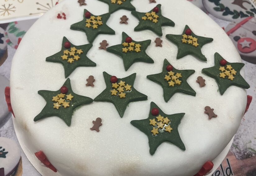 Festive cake topped with green star decorations and small brown figures on a white icing base. The tone is celebratory and joyful.