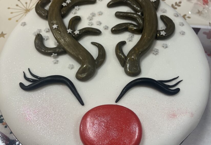 Festive cake decorated as a reindeer face with chocolate antlers, closed eyes with lashes, and a shiny red nose. Background includes party elements.