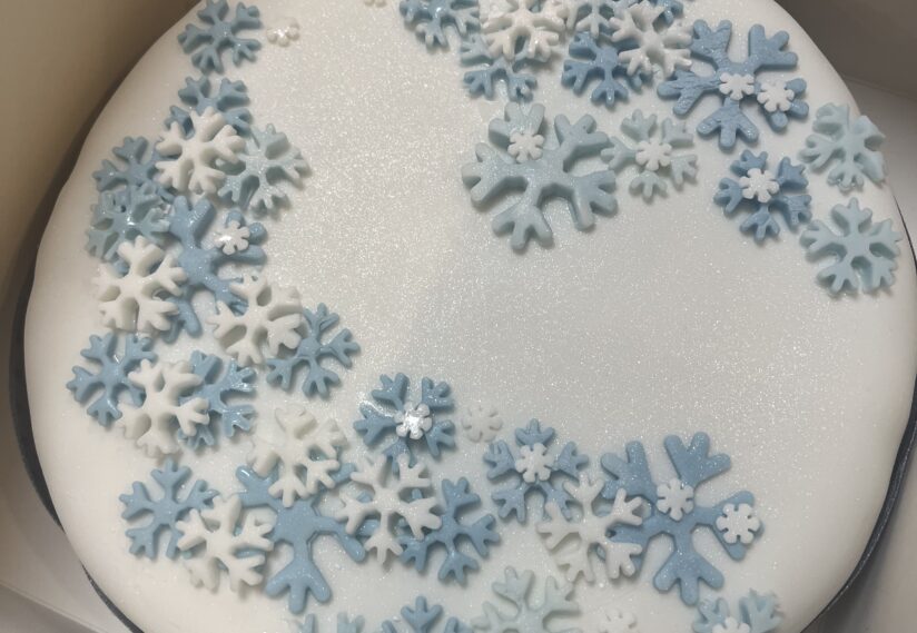 Circular cake with white icing and blue and white snowflake decorations, giving a wintry feel. The cake surface has a subtle shimmer.