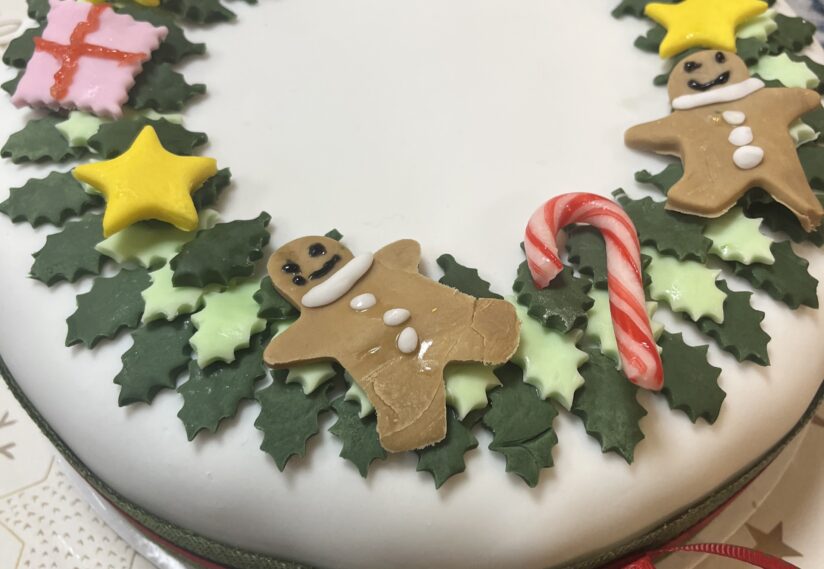 Festive cake with white icing, decorated with gingerbread men, candy canes, yellow stars, pink gift boxes, and green holly leaves, evoking holiday cheer.