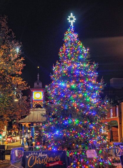 A festive crowd gathers at night around a brightly lit Christmas tree, adorned with colorful lights.