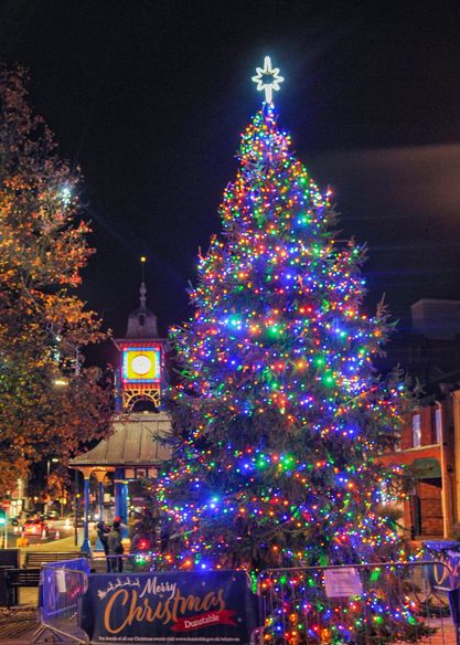 A festive crowd gathers at night around a brightly lit Christmas tree, adorned with colorful lights.