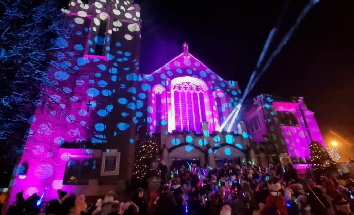 A church facade illuminated with colorful lights. Festive atmosphere with crowds gathered, conveying holiday cheer.