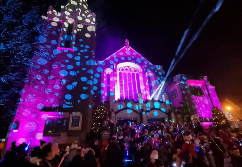 A church facade illuminated with colorful lights. Festive atmosphere with crowds gathered, conveying holiday cheer.