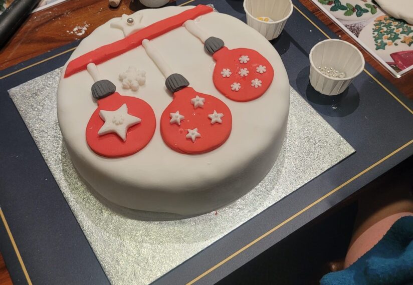 A festively decorated cake with white icing and red fondant baubles, adorned with stars and snowflakes. Baking tools and ingredients surround it.