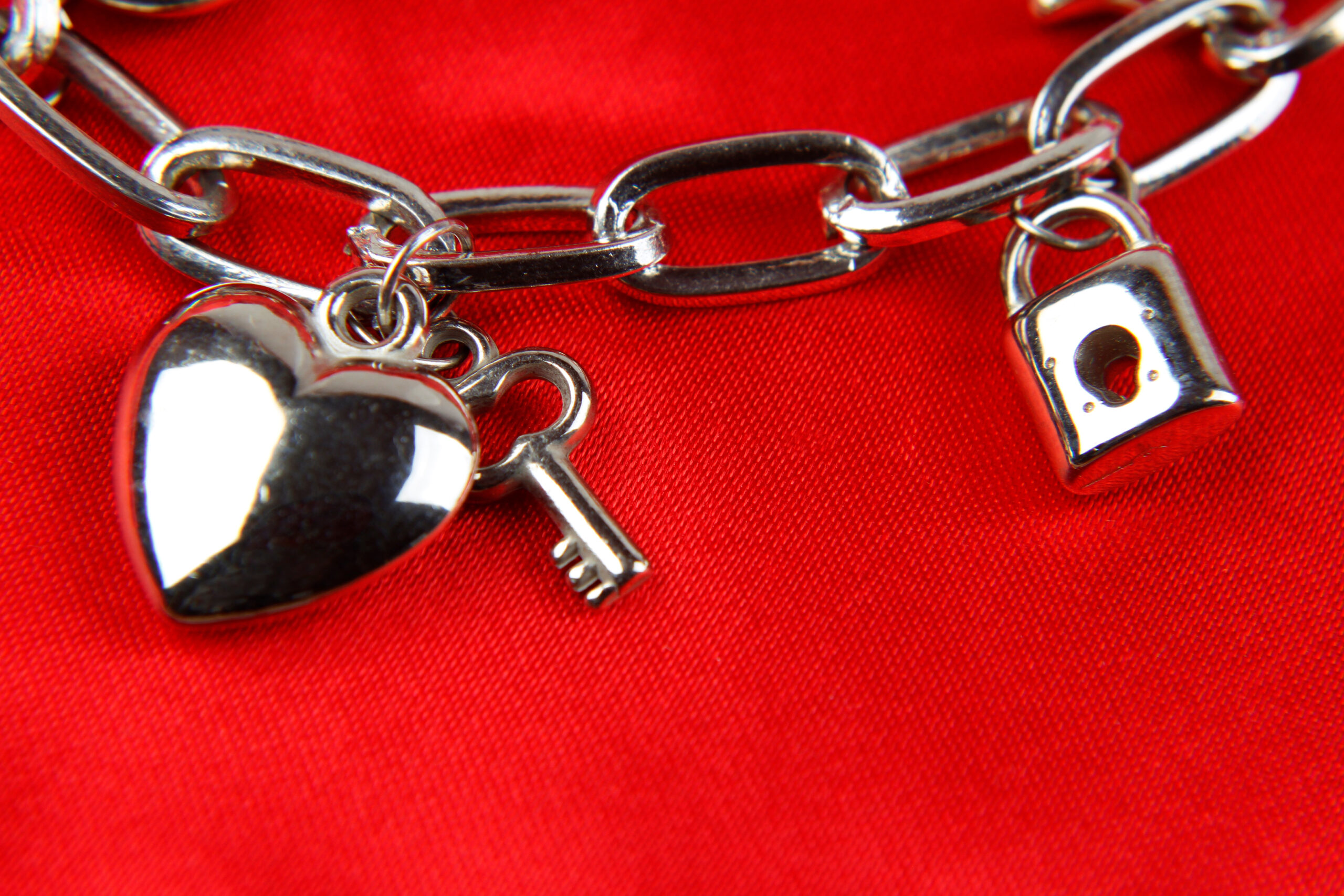 Silver charm bracelet on vibrant red background, featuring a heart, key, and padlock. Reflective, romantic, and elegant theme.