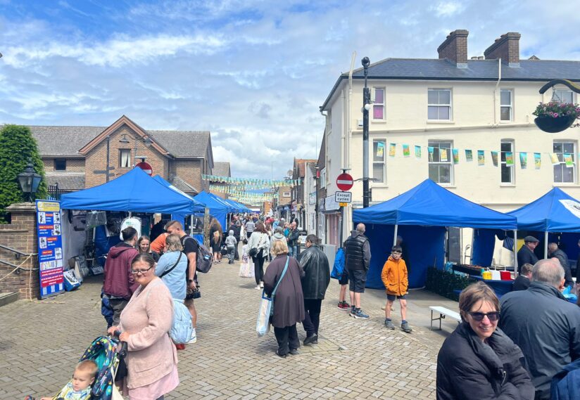 Street market scene with blue tents lining a cobblestone road. People stroll and browse stalls under a partly cloudy sky, creating a lively, vibrant atmosphere.