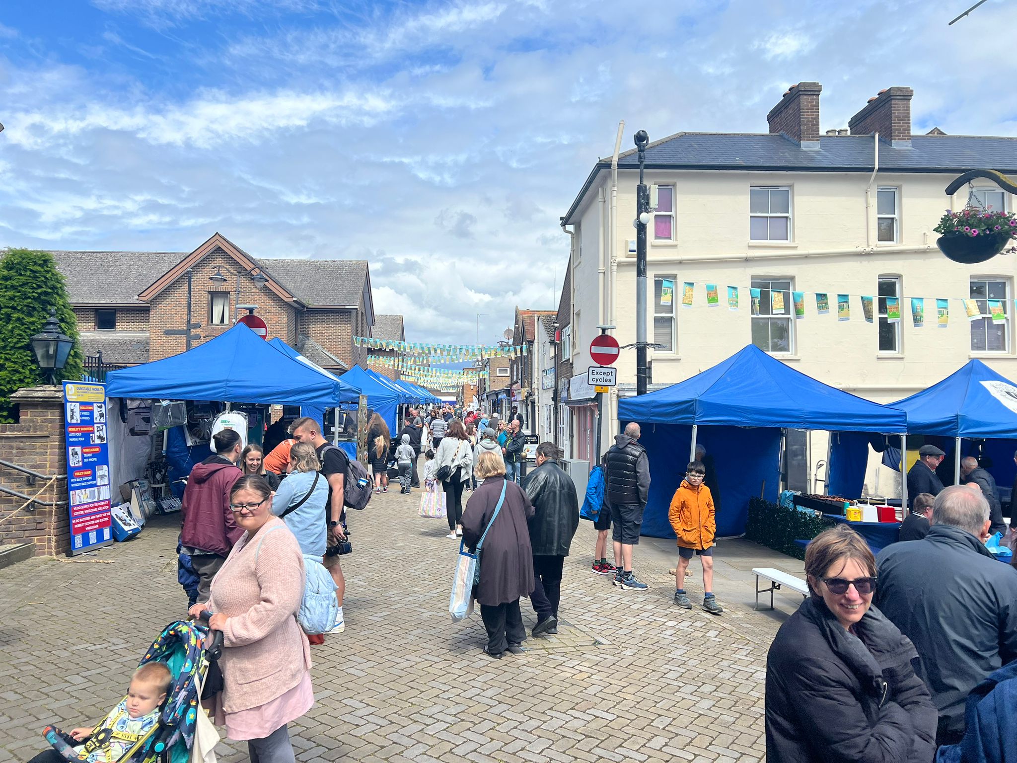 Street market scene with blue tents lining a cobblestone road. People stroll and browse stalls under a partly cloudy sky, creating a lively, vibrant atmosphere.