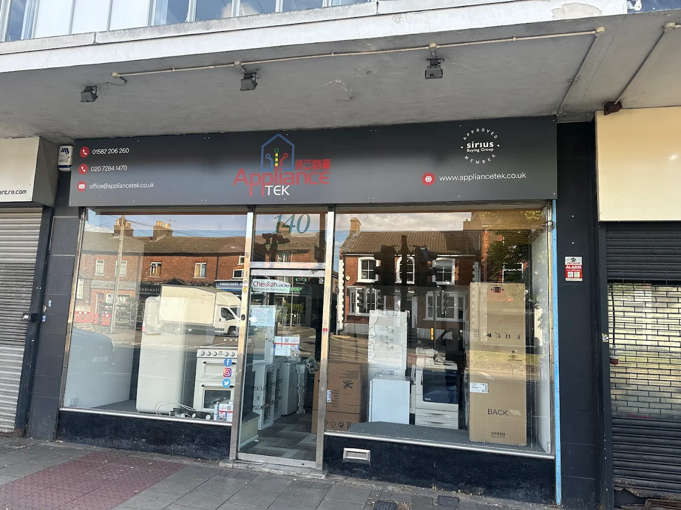 Storefront of an appliance shop with large glass windows, showing boxes and washing machines inside. Signs and contact info displayed on the black banner.