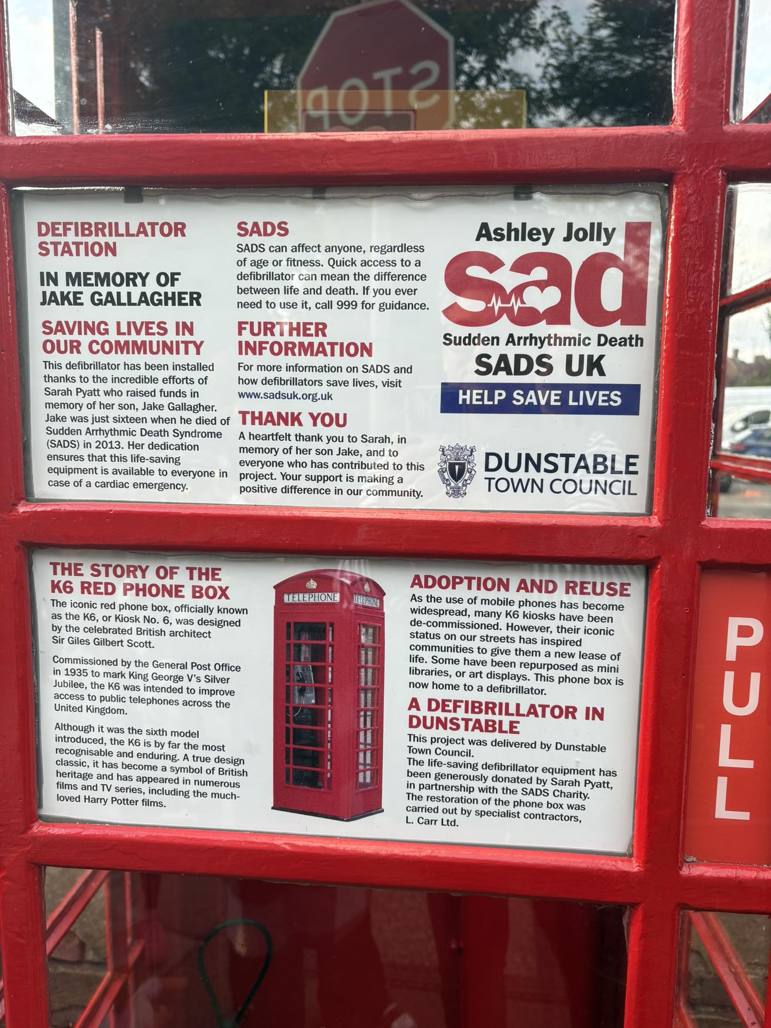 Red phone box converted into a defibrillator station with informational posters about SADS, defibrillator use, and the history and reuse of the K6 phone box.