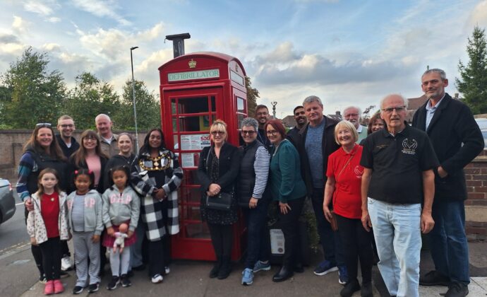 A diverse group of people gathered around a red telephone booth converted into a defibrillator station against a cloudy sky.