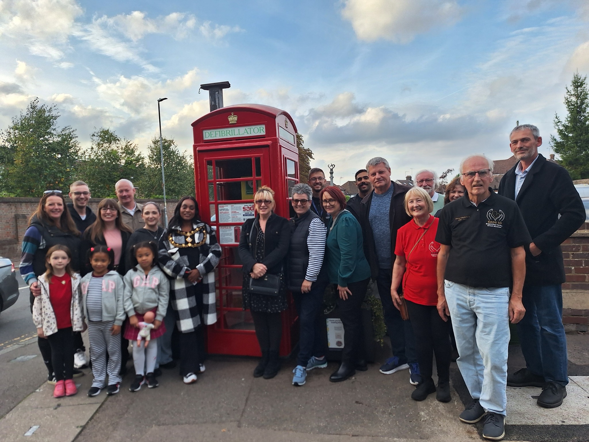 A diverse group of people gathered around a red telephone booth converted into a defibrillator station against a cloudy sky.