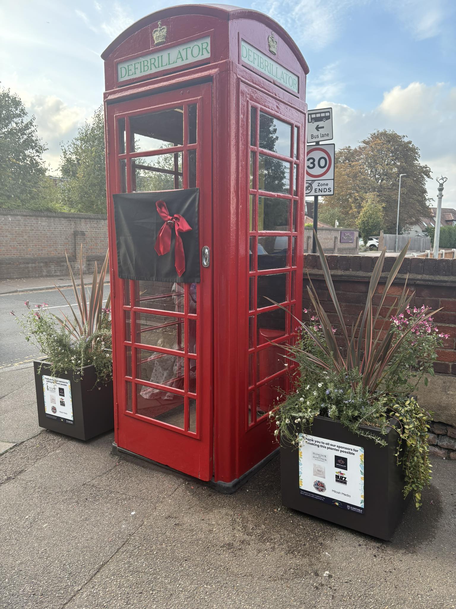 A red phone box repurposed as a defibrillator station, decorated with a black cloth and red ribbon, stands between two flower planters on a paved sidewalk.