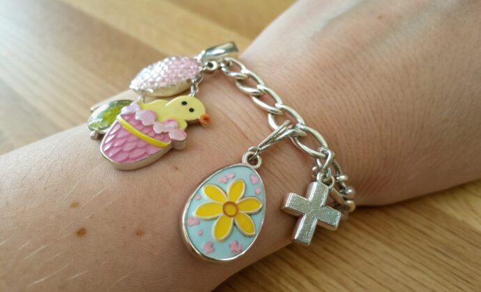 A silver charm bracelet with a cross, a flower-decorated egg, a yellow chick, and a pink gem charm is worn on a wrist over a wooden surface.