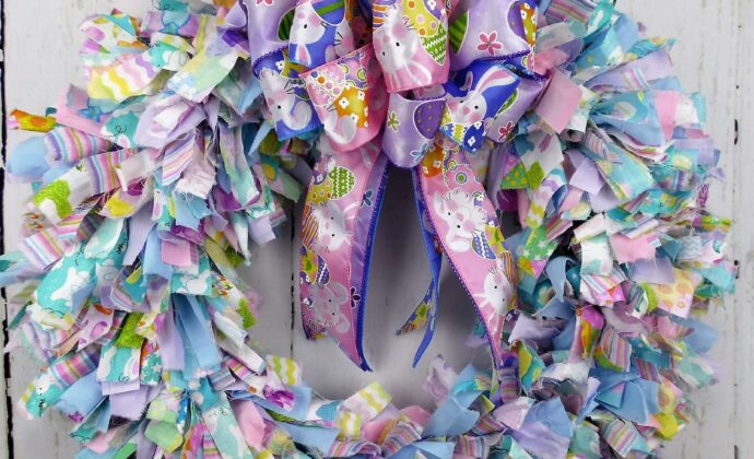 Colorful Easter wreath with pastel fabric strips and a large purple bow with bunny motifs. Vibrant and festive against a rustic white wooden background.