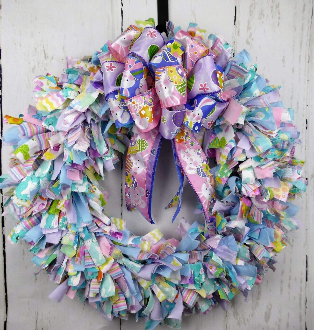 Colorful Easter wreath with pastel fabric strips and a large purple bow with bunny motifs. Vibrant and festive against a rustic white wooden background.