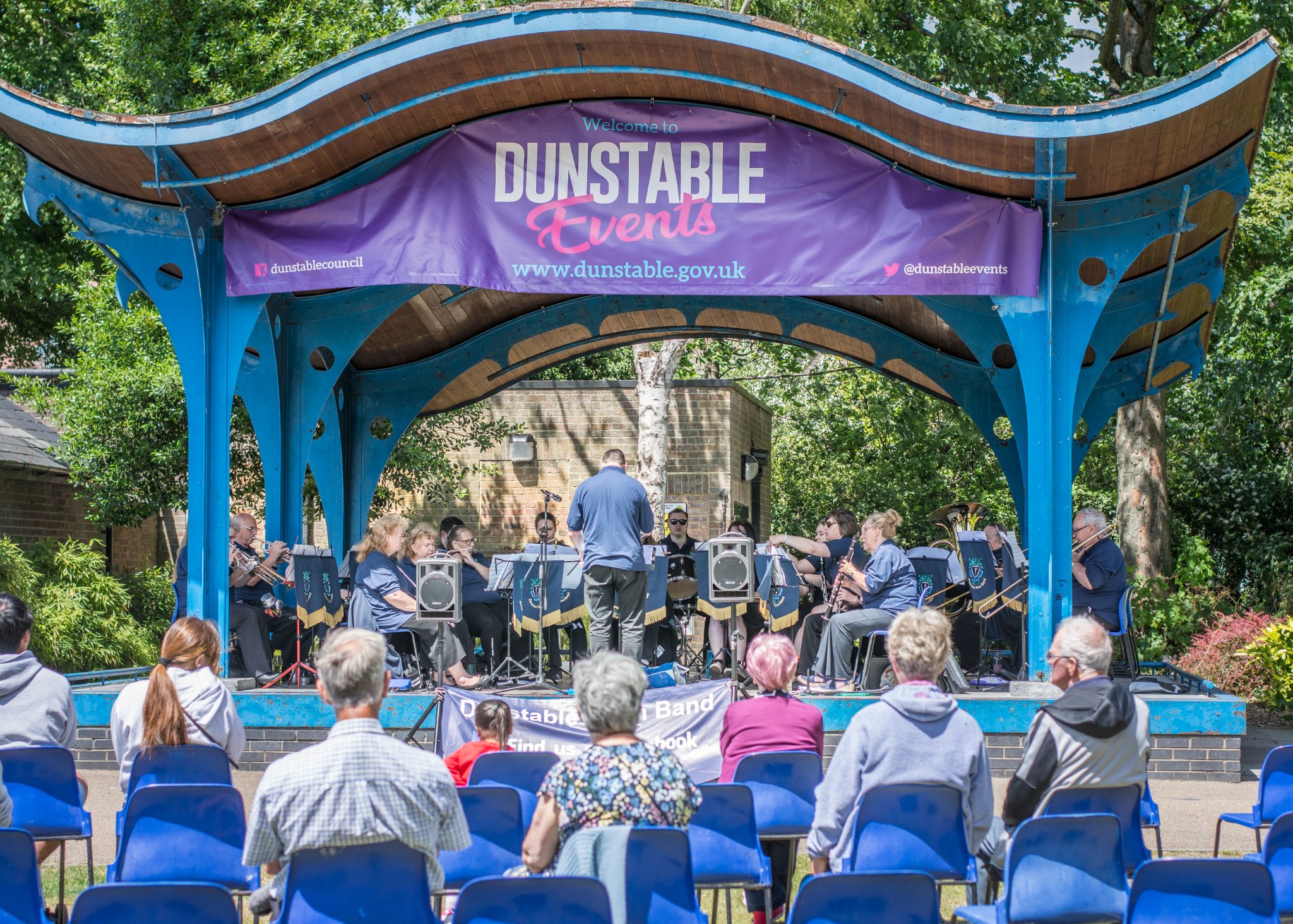 Outdoor concert with a band playing on a blue stage under a "Dunstable Events" banner. Audience seated in front, enjoying a sunny day.
