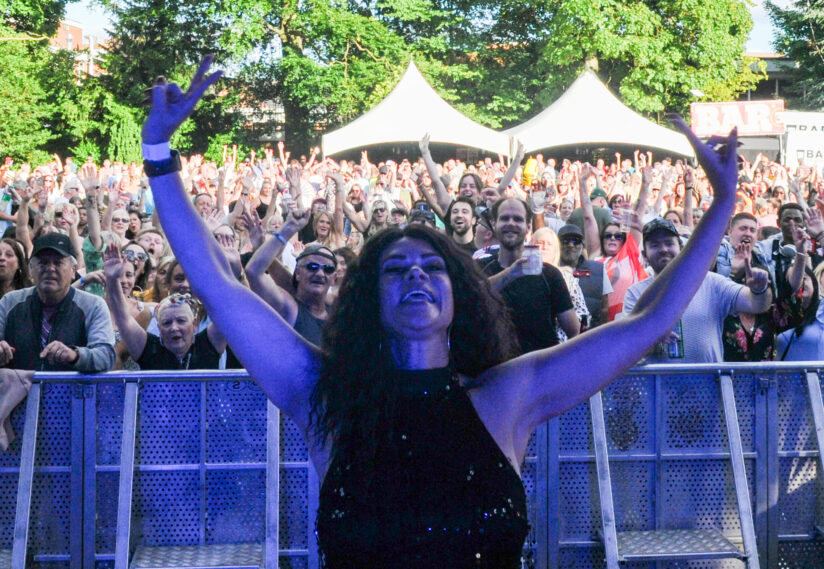 A woman joyfully raises her arms at a lively outdoor concert, facing a cheering crowd. Tents and trees are visible in the background. Festive atmosphere.