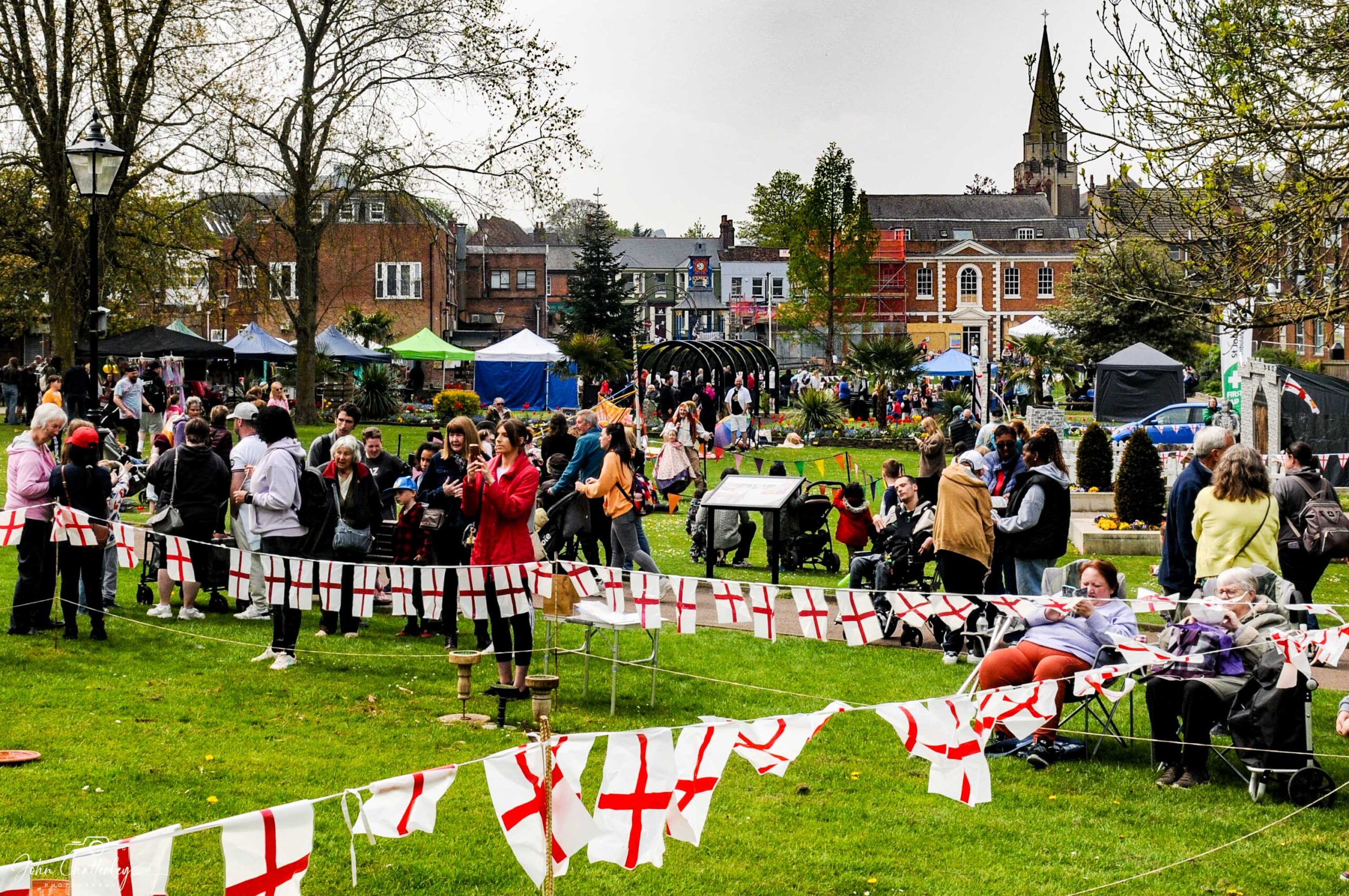 A lively outdoor festival with people gathered on grass, surrounded by red-and-white St. George's flags. Tents and buildings are visible in the background.