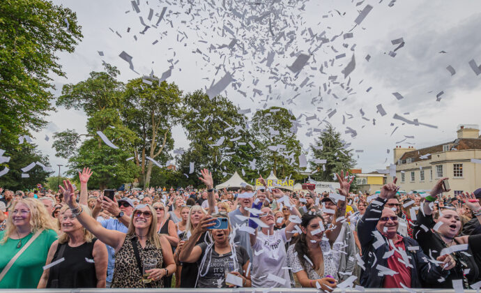 A lively crowd at an outdoor concert, with people cheering and raising their hands as white confetti fills the sky. Trees and a building are in the background.