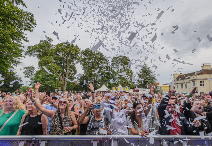 A lively crowd at an outdoor concert, with people cheering and raising their hands as white confetti fills the sky. Trees and a building are in the background.