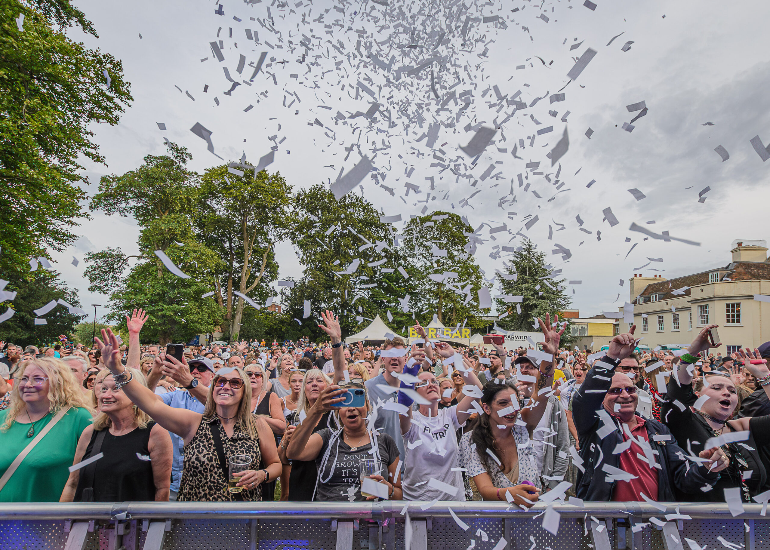 A lively crowd at an outdoor concert, with people cheering and raising their hands as white confetti fills the sky. Trees and a building are in the background.