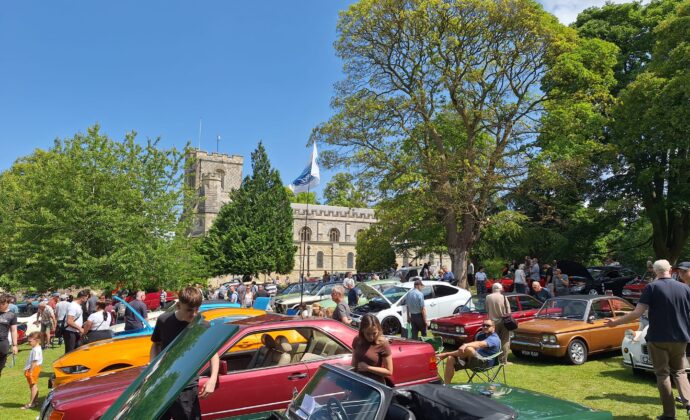 A vibrant car show on a sunny day features classic cars parked on a green lawn. People admire the vehicles under a clear blue sky, with a church in the background.