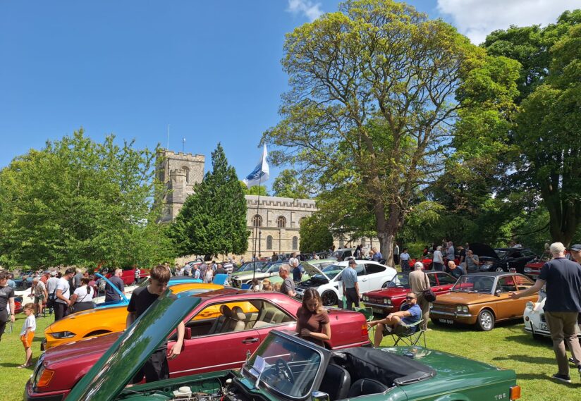 A vibrant car show on a sunny day features classic cars parked on a green lawn. People admire the vehicles under a clear blue sky, with a church in the background.