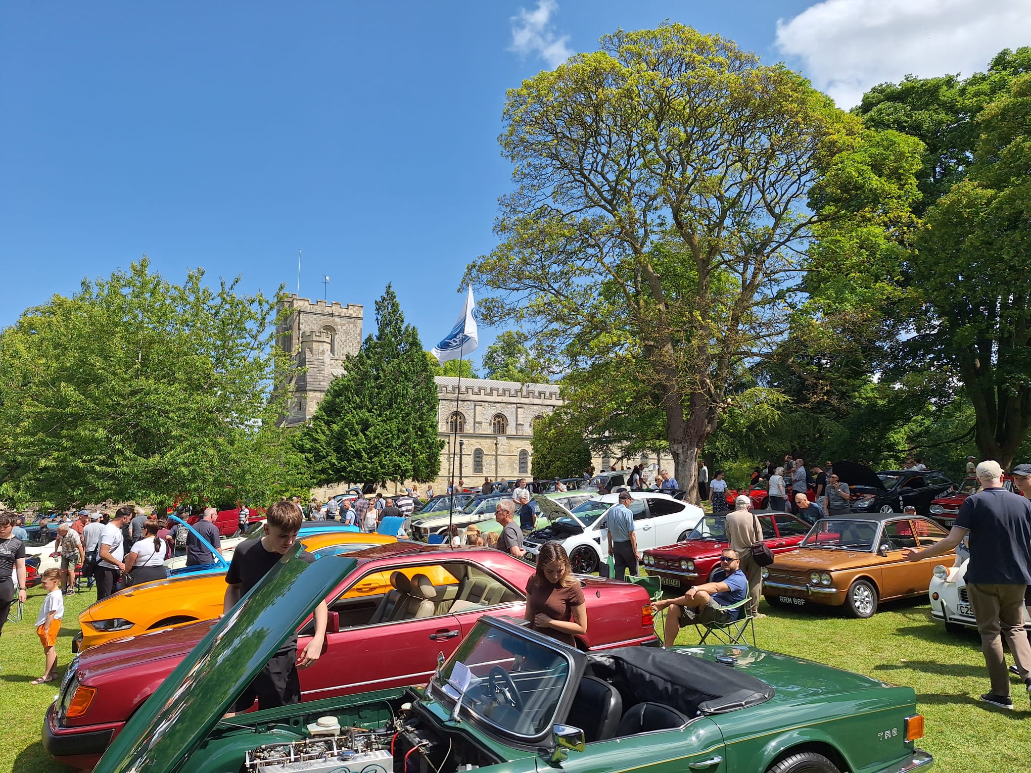 A vibrant car show on a sunny day features classic cars parked on a green lawn. People admire the vehicles under a clear blue sky, with a church in the background.