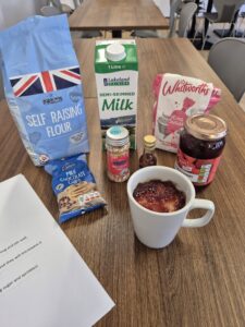 Various baking ingredients, including flour, milk, sugar, chocolate chips, sprinkles, vanilla extract, and jam, are arranged on a wooden table next to a mug with jam on top.