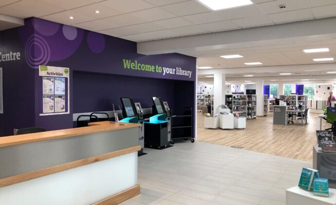 Modern library interior with a wooden reception desk, self-service machines, and bookshelves. The sign reads 