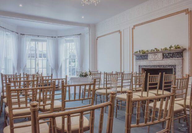 Elegant small wedding venue with gold chairs facing a fireplace adorned with greenery. Sunlight streams through sheer white curtains, creating a serene ambiance.