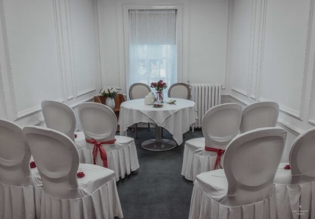 Small, elegant room with white draped chairs arranged in rows. A round table with floral centerpieces stands by a curtained window, creating a serene atmosphere.