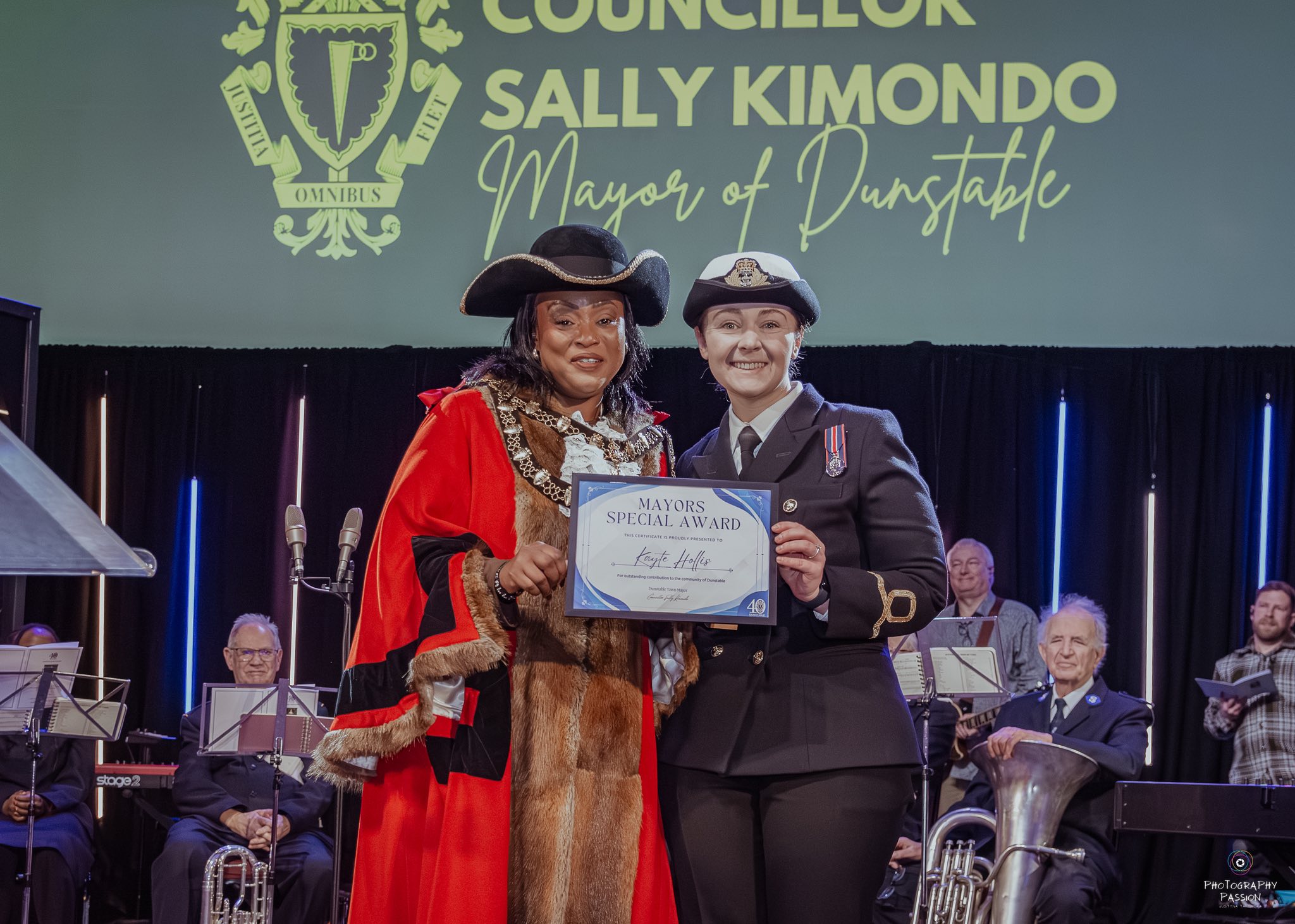 Two people stand smiling, one in a red ceremonial robe and the other in a uniform, holding a 