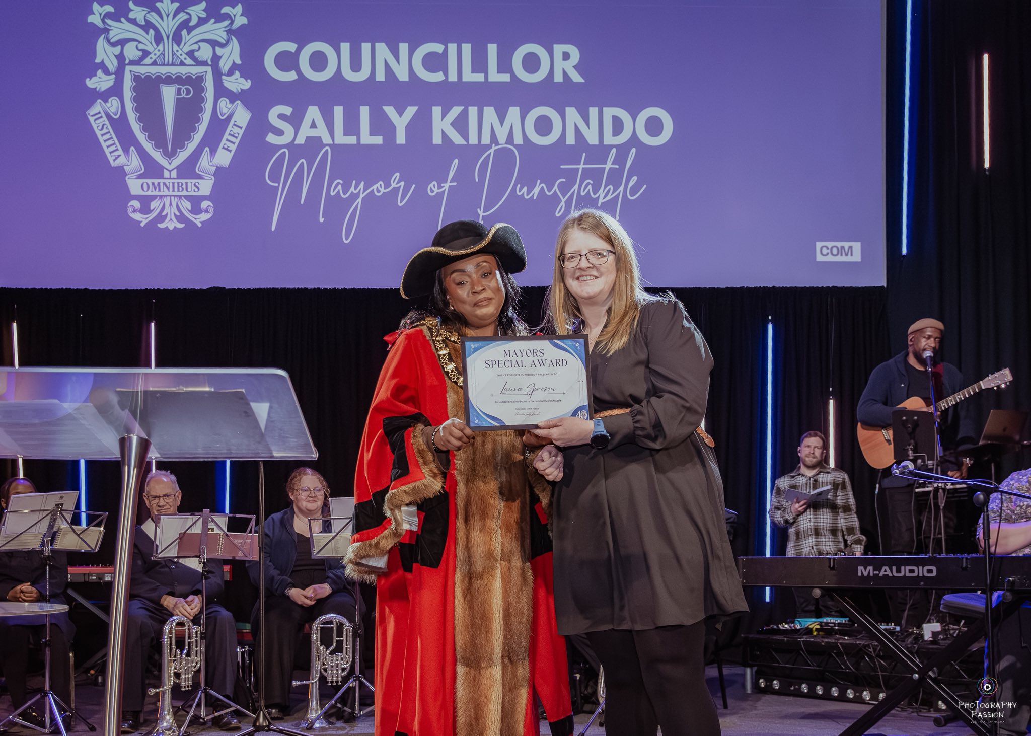 Mayor in ceremonial robes presents a special award to a smiling woman on stage. A band and audience are in the background, under a spotlight.