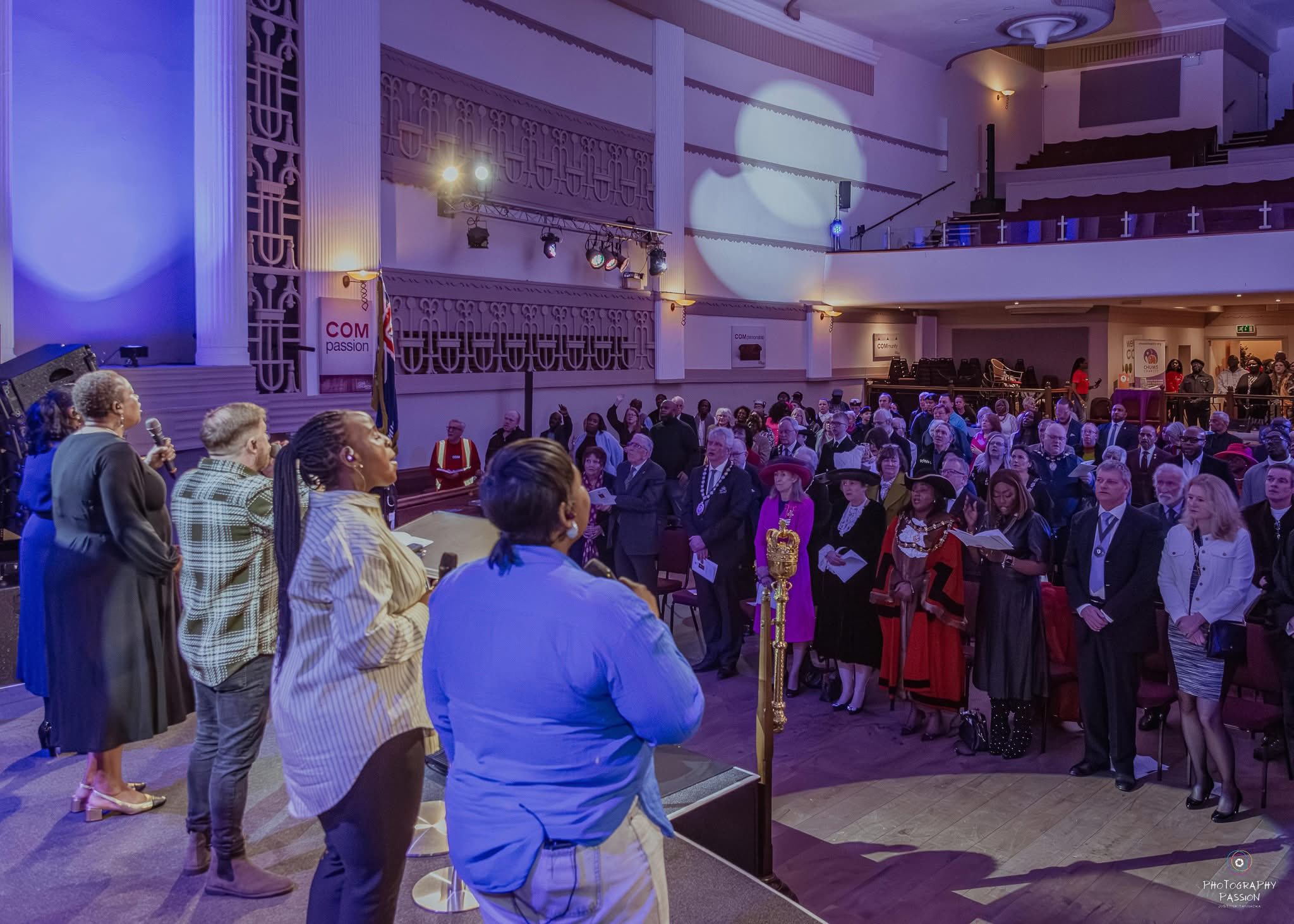 A group of singers performs on stage facing an audience in a grand hall. The crowd, including people in formal attire and hats, watches attentively. Blue lighting creates a vibrant atmosphere.