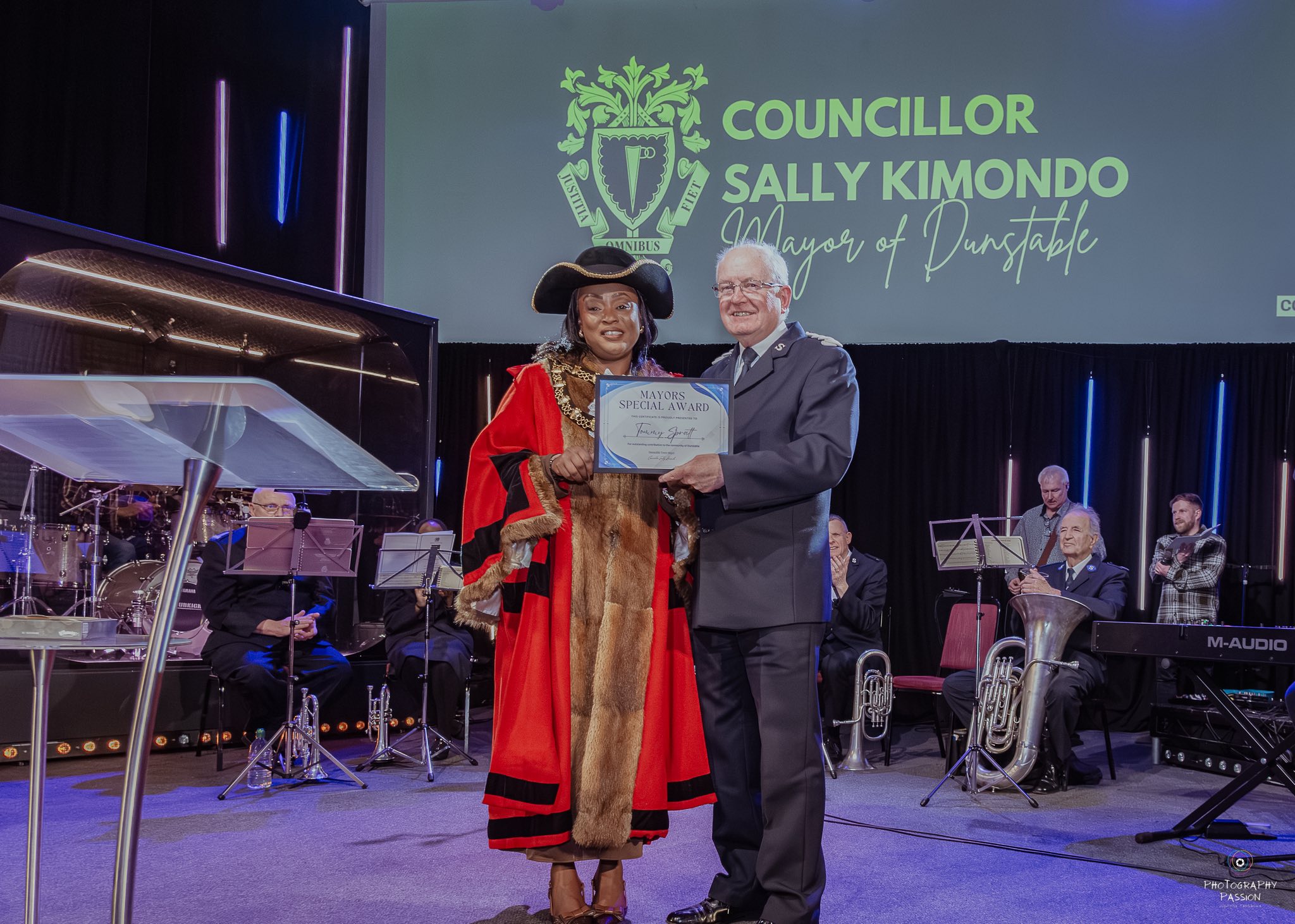 Councillor in ceremonial attire presents an award to a man on stage. Background shows musicians and large sign with her name and title. Festive and formal.