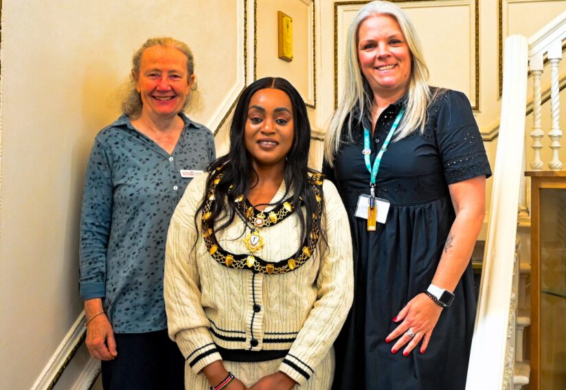 Three women stand side by side on a staircase, smiling. The central woman wears a ceremonial chain, suggesting a formal occasion. Warm atmosphere.